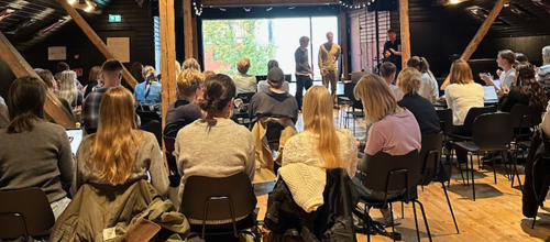 Folk sidder på stole i et lokale og lytter til tre personer, der står foran et stort vindue og taler.